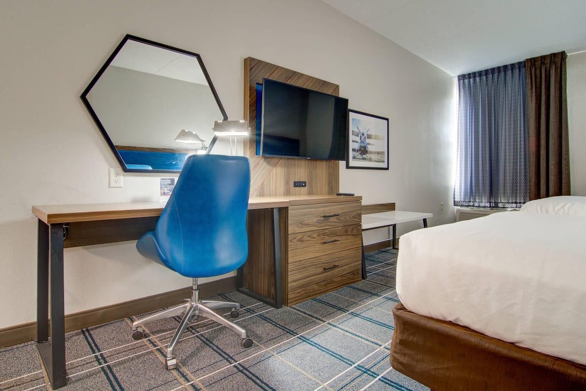 Een hotelkamer met een bed, een bureau met stoel, een televisie, een spiegel en een raam met gordijnen.