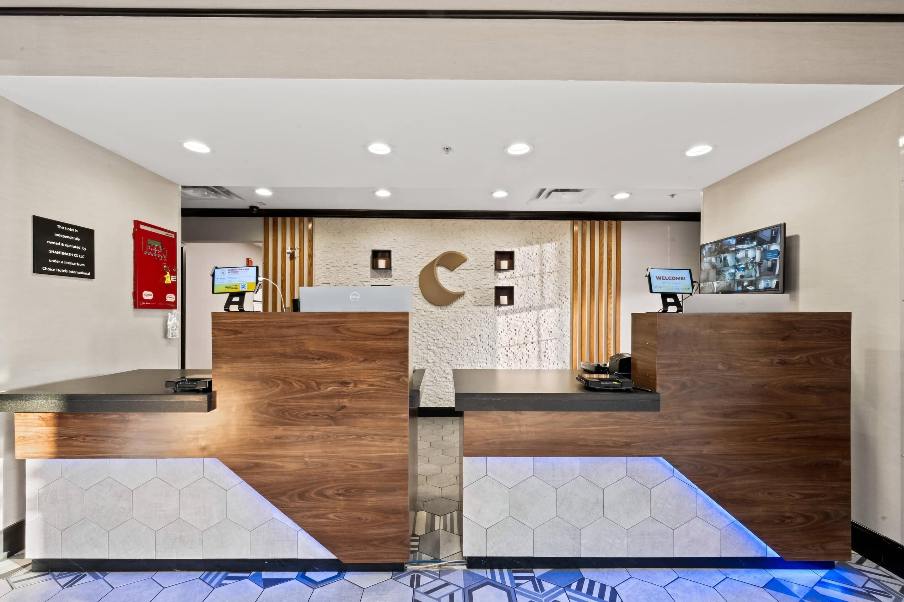 A reception area with wooden reception desks, a digital check-in kiosk, a fire alarm, and a welcome sign.