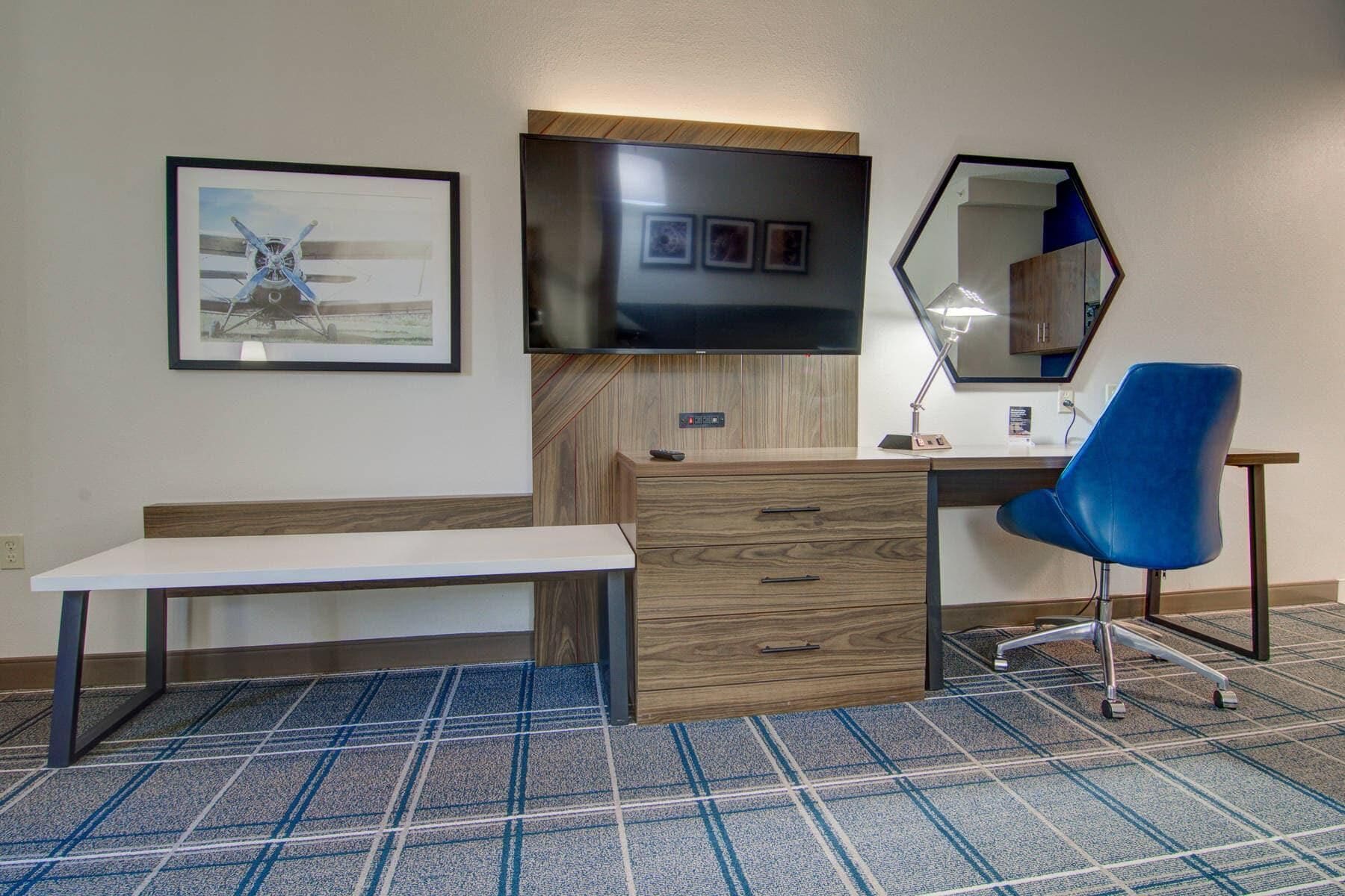 Habitación de hotel con un escritorio de madera, una silla azul, un banco, un televisor de pantalla plana, un espejo y un cuadro en la pared.