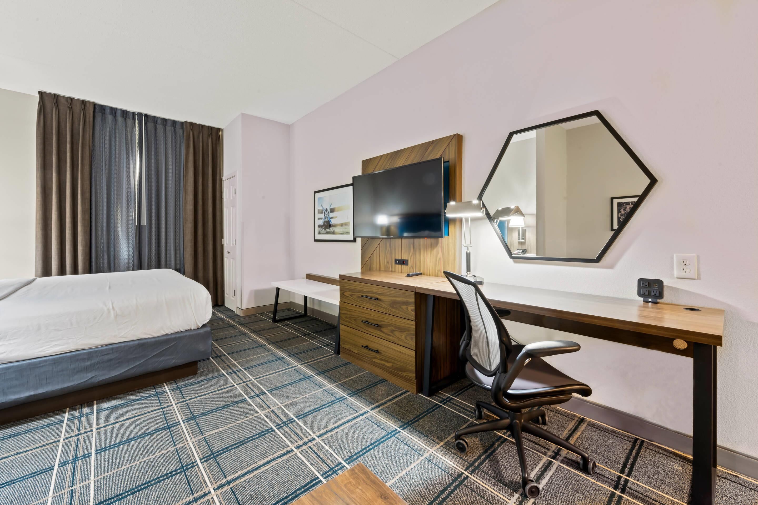 Una habitación de hotel con cama, escritorio, computadora, silla, televisor y espejo.