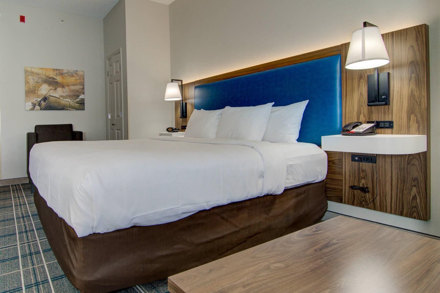 Een hotelkamer met een groot bed, een houten hoofdbord, een nachtkastje met een lamp, een klein bureau met een telefoon en een schilderij aan de muur.