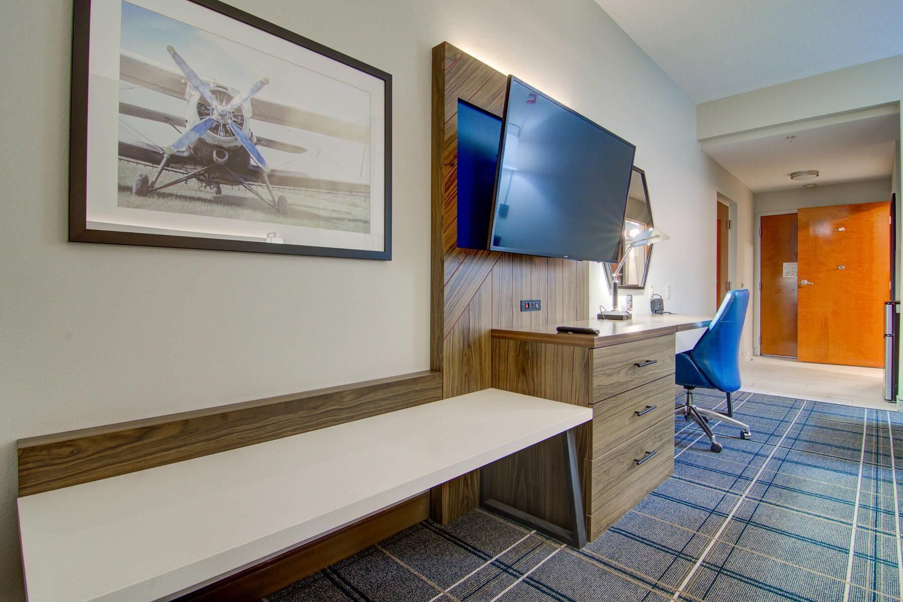 Habitación de hotel con un banco de madera, un escritorio con silla, un televisor de pantalla plana en la pared y un cuadro enmarcado de un avión.