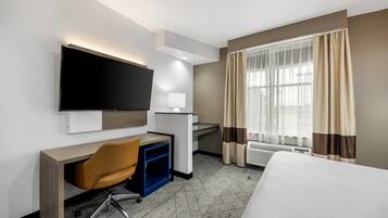 Habitación de hotel con cama, escritorio, silla, televisor y ventana con cortinas.