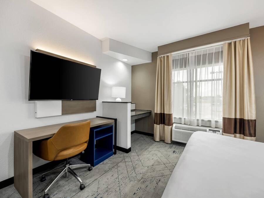 Habitación de hotel con cama, escritorio, silla, televisor y ventana con cortinas.