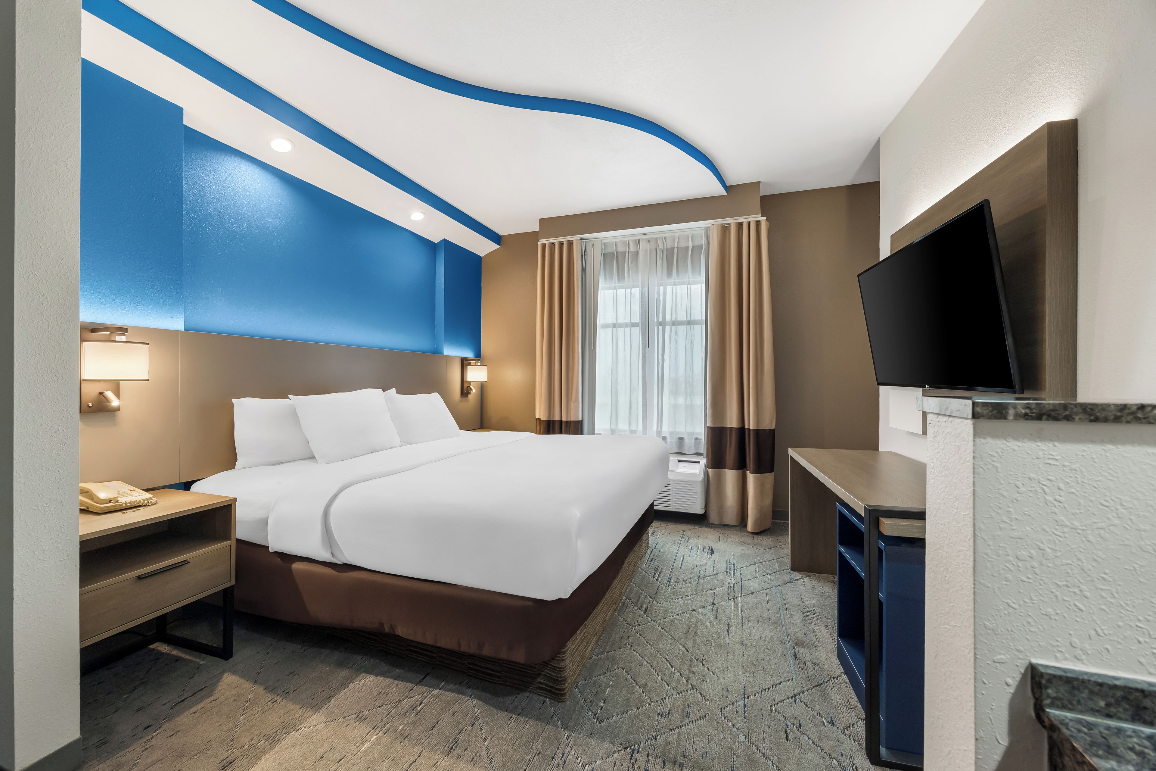 Une chambre d’hôtel comprenant un lit, des tables de chevet, un bureau, un téléviseur à écran plat et une fenêtre avec des rideaux.