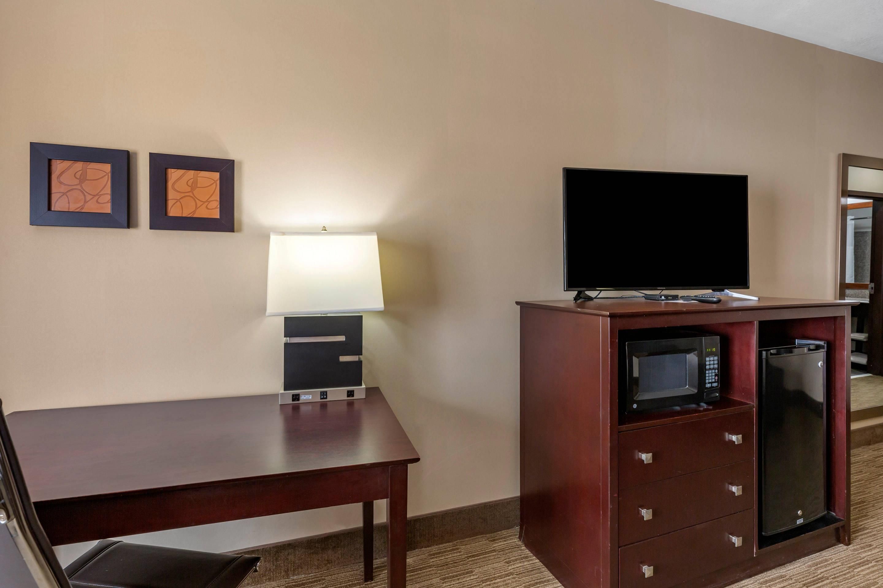 A hotel room with a wooden desk, a television on a stand, a microwave, and a trash bin.