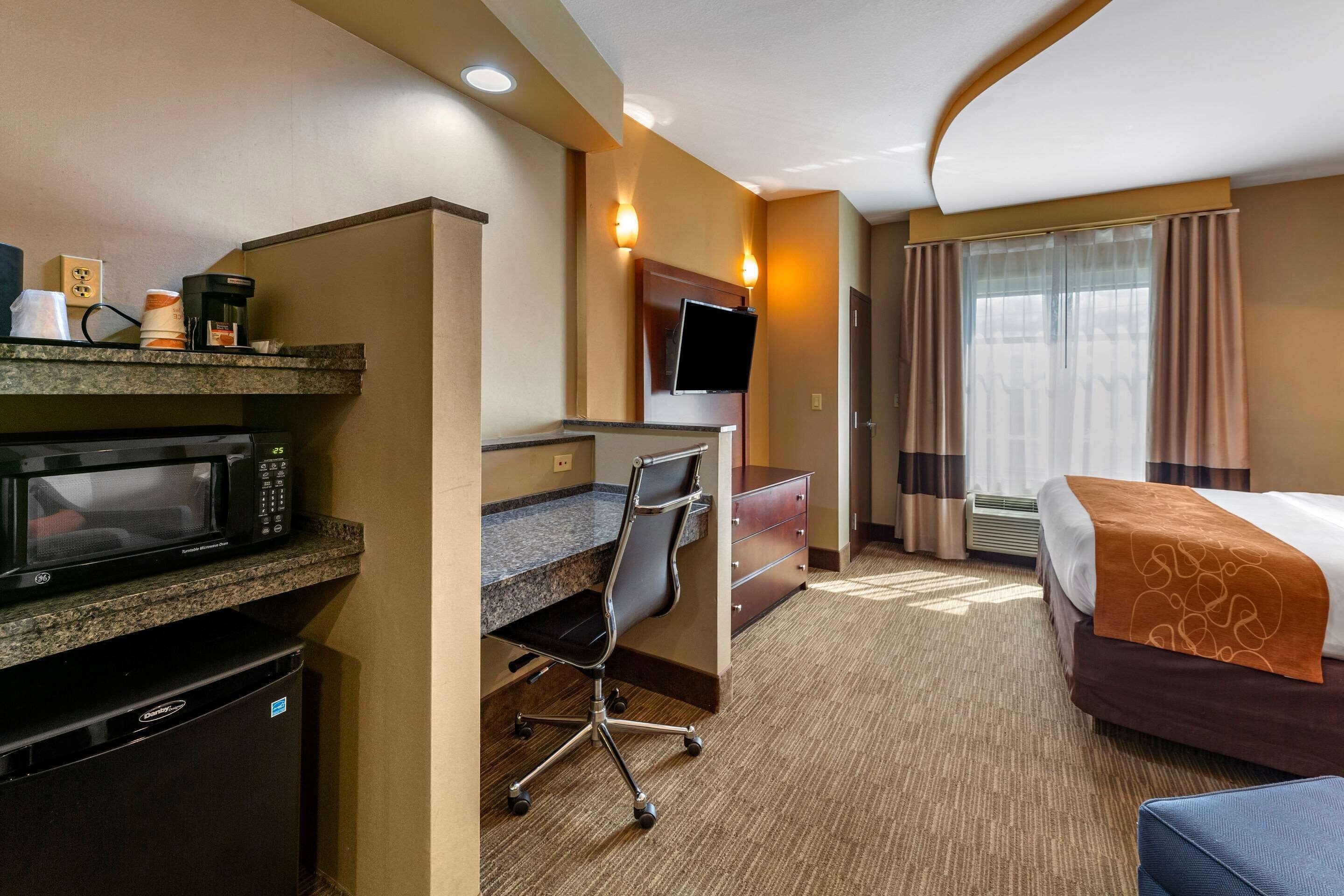 Habitación de hotel con kitchenette, un escritorio y una silla, una cama con cabecera marrón y un televisor montado en la pared.
