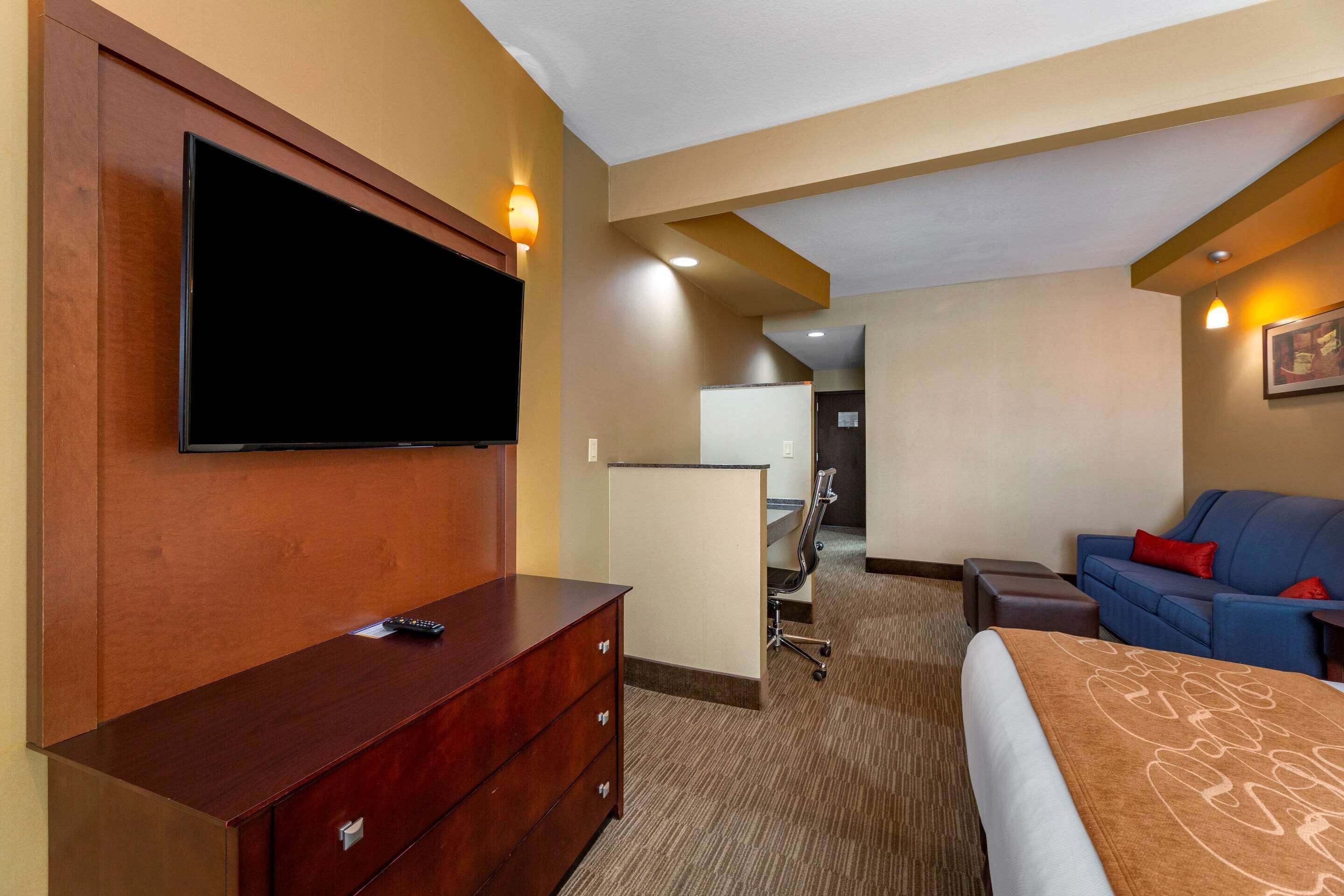 Habitación de hotel con televisor de pantalla plana montado en la pared, cómoda de madera, sofá azul con cojines rojos, escritorio con silla y piso alfombrado.