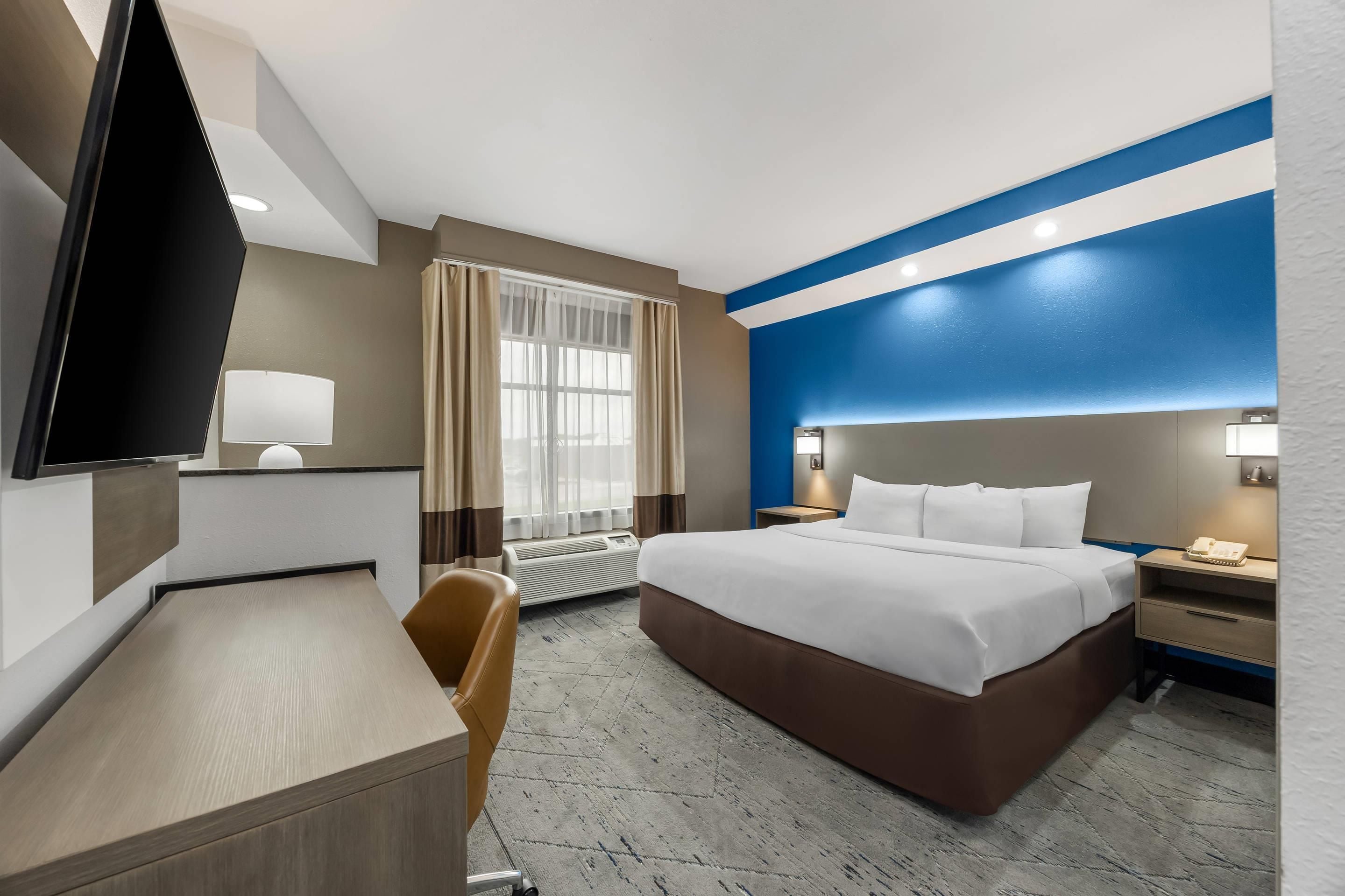 Habitación de hotel con cama, escritorio, silla, televisor y un amplio ventanal con cortinas.