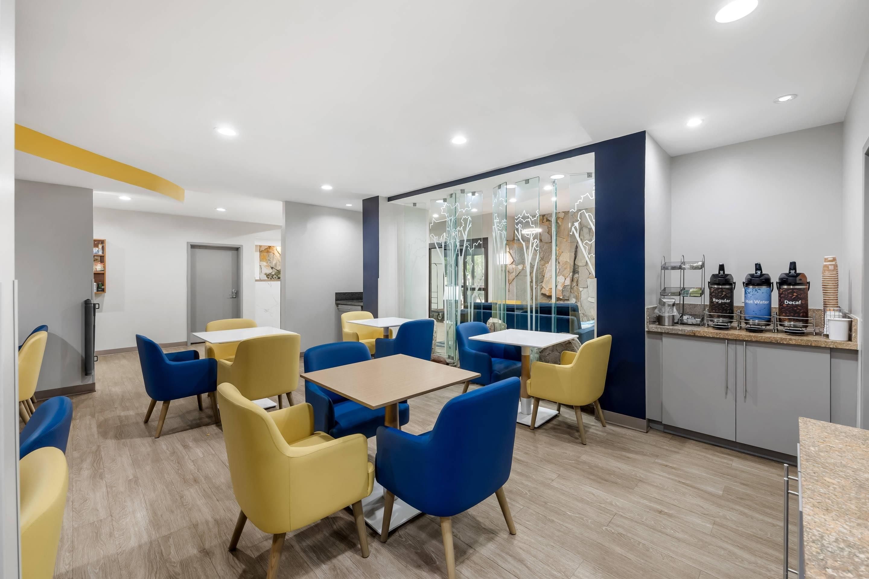 Un café moderne avec des chaises jaunes et bleues, un comptoir où sont servis des boissons et un mur en pierre orné d’œuvres d’art abstraites.