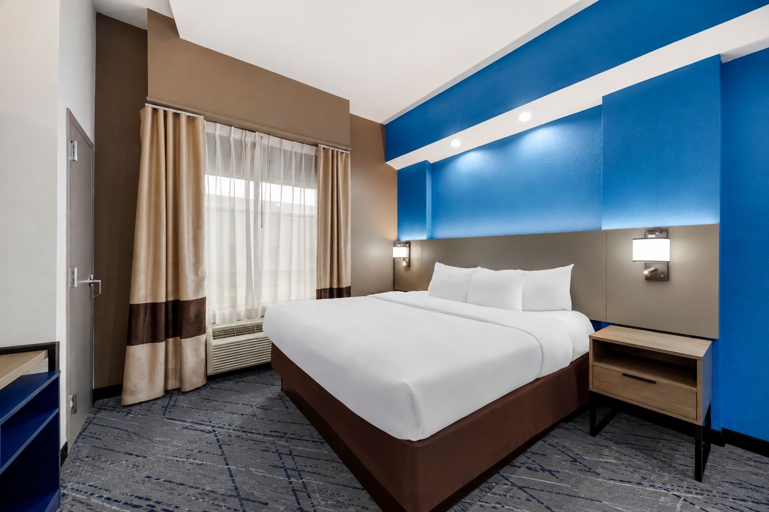 Una habitación de hotel con una cama grande, una mesita de noche, una lámpara de pared y un ventanal con cortinas.