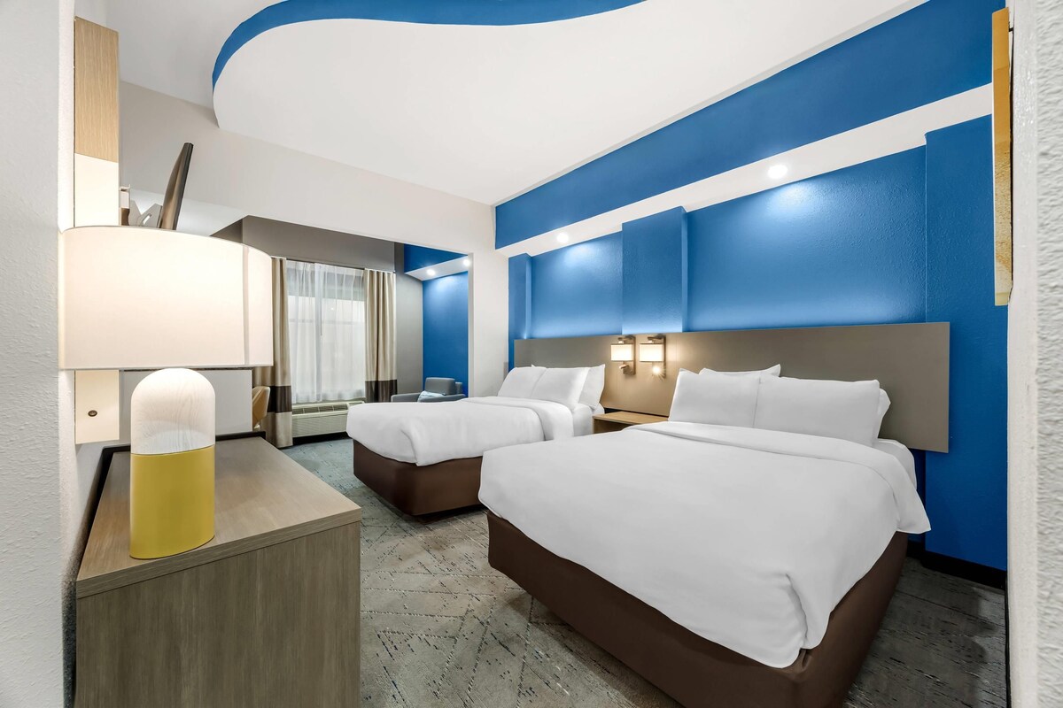 Ein Hotelzimmer mit zwei Betten, einem Schreibtisch mit Lampe und einem großen Fenster mit Vorhängen.