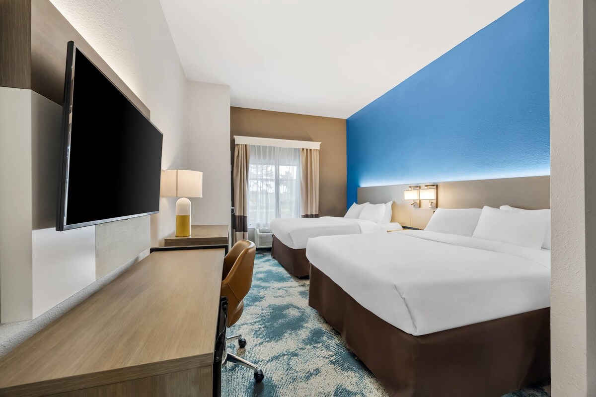 Ein Hotelzimmer mit Flachbildfernseher, Schreibtisch, zwei Betten, einem Sessel und einem großen Fenster mit Vorhängen.