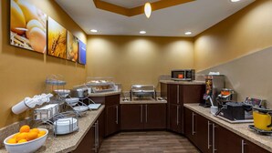 A hotel breakfast area with a buffet setup, including a bowl of oranges, a microwave, and various breakfast items on the counter.