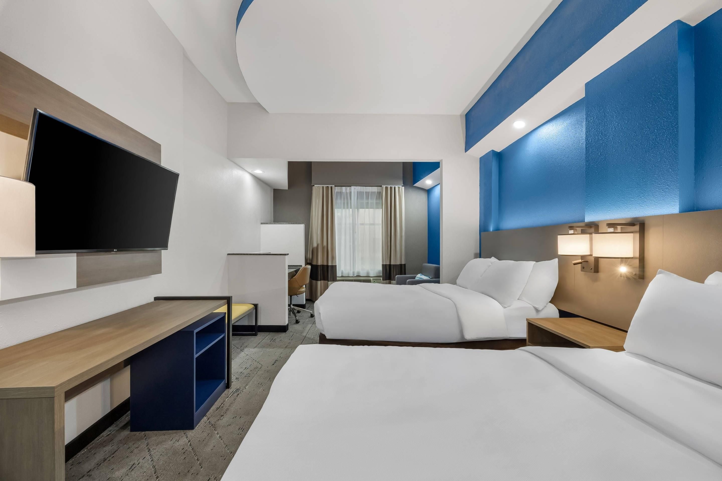A hotel room with a large flat-screen TV, two beds, a desk, and a seating area.