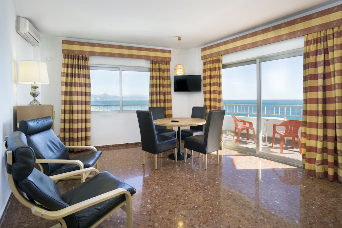 Une pièce avec un rideau à carreaux, une table ronde entourée de chaises, un téléviseur et une vue sur la mer.