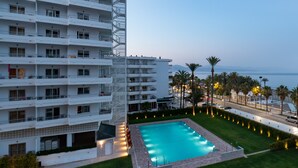 Un hotel de varios pisos con piscina, rodeado de palmeras y vista al mar.