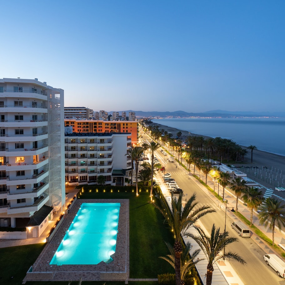 A hotel with a swimming pool, beachfront view, and a road with palm trees.