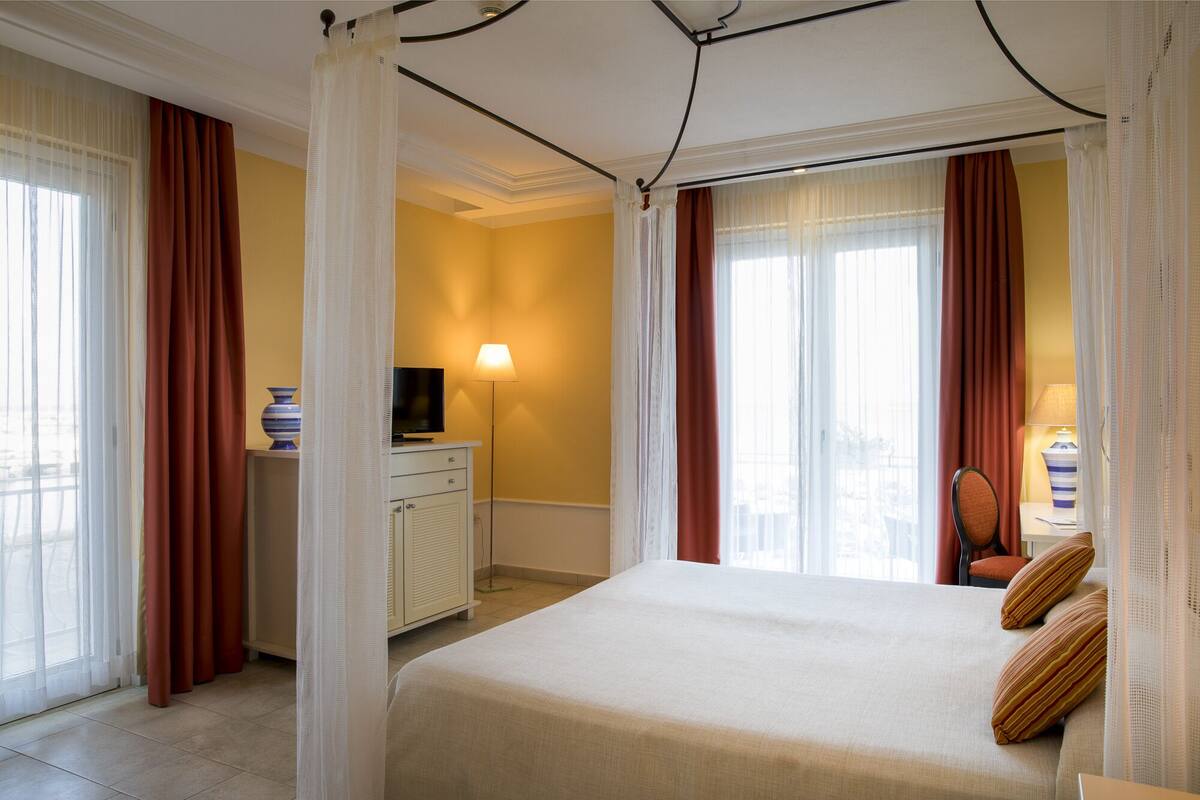 Ein Hotelzimmer mit Bett, Schreibtisch mit Fernseher, Lampe und Blick auf die Stadt durch große Fenster.