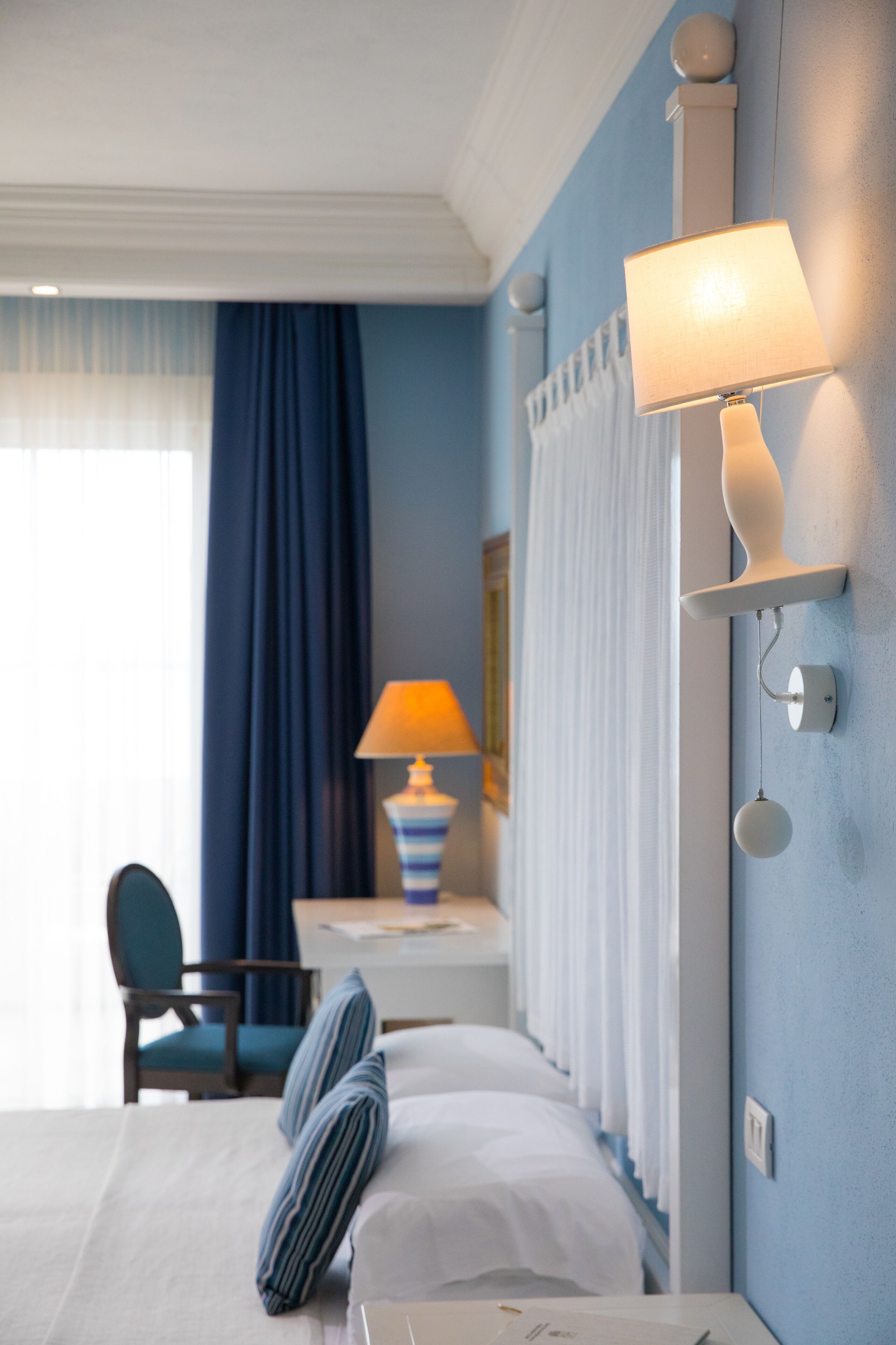 A hotel room with a bed, a desk with a lamp, a chair, and a wall-mounted lamp.