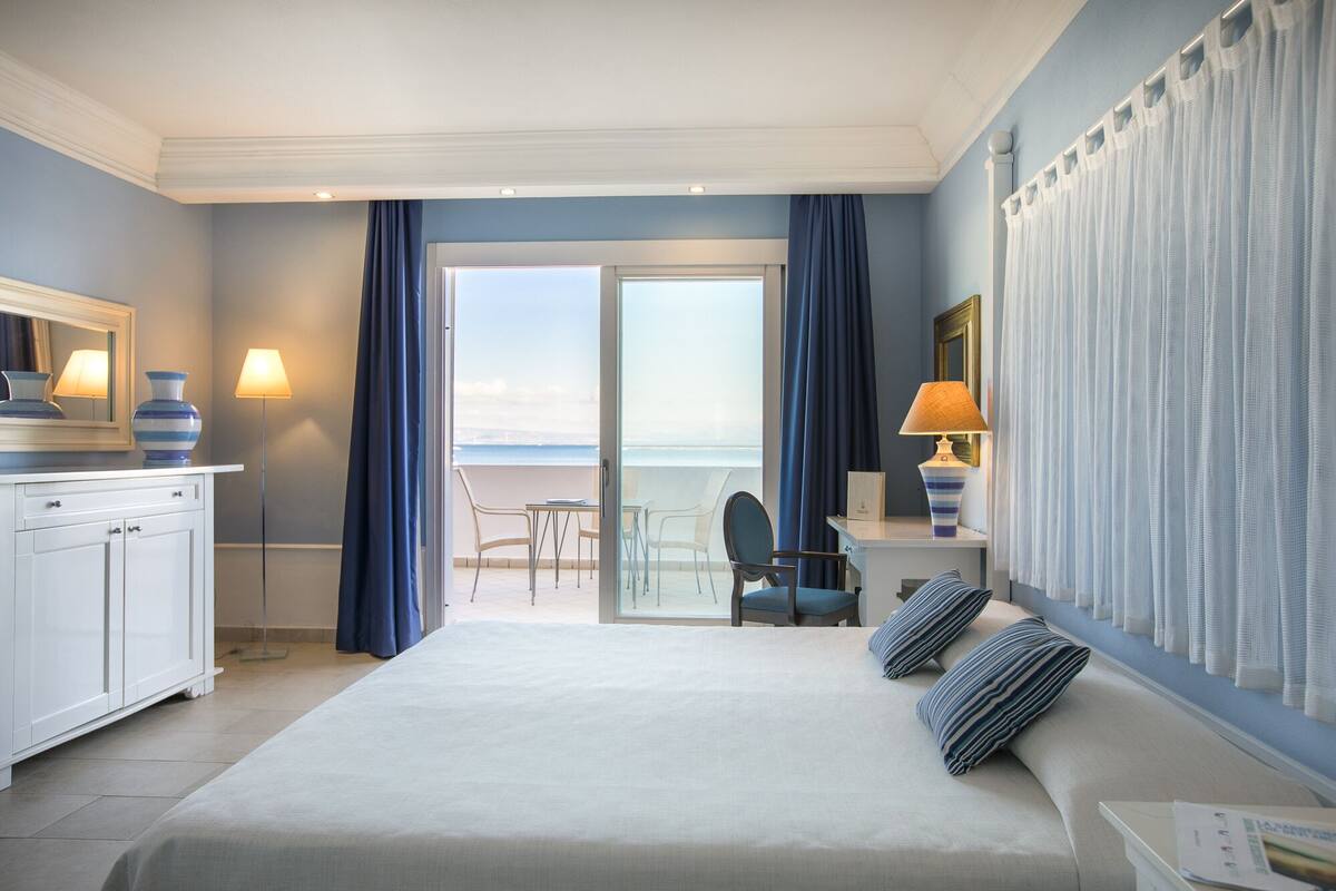 Ein Hotelzimmer mit Bett, Schreibtisch mit Lampe, Stuhl und Meerblick durch eine Glastür.
