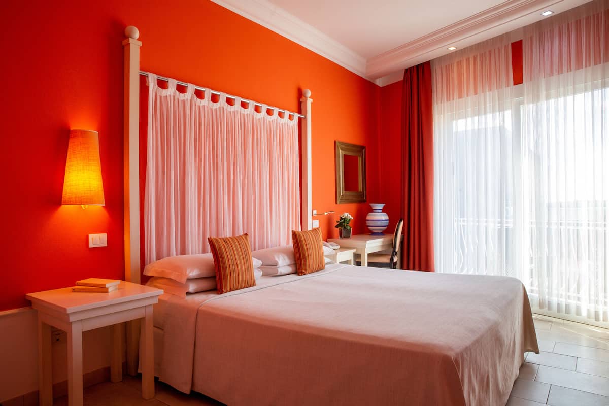 Ein Hotelzimmer mit einem Himmelbett, einem Nachttisch mit Lampe, einem Schreibtisch mit Stuhl und einem großen Fenster mit durchsichtigen Vorhängen.