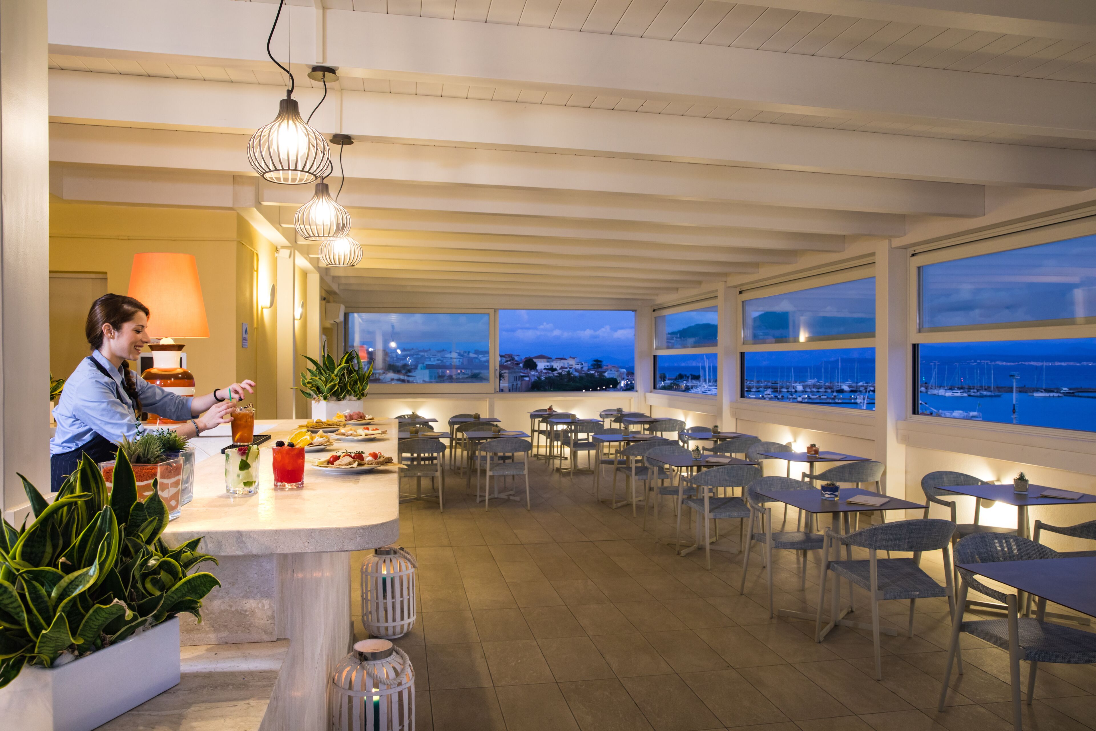 A restaurant with a view of the marina, tables and chairs arranged for dining, and a bar area with a person preparing drinks.
