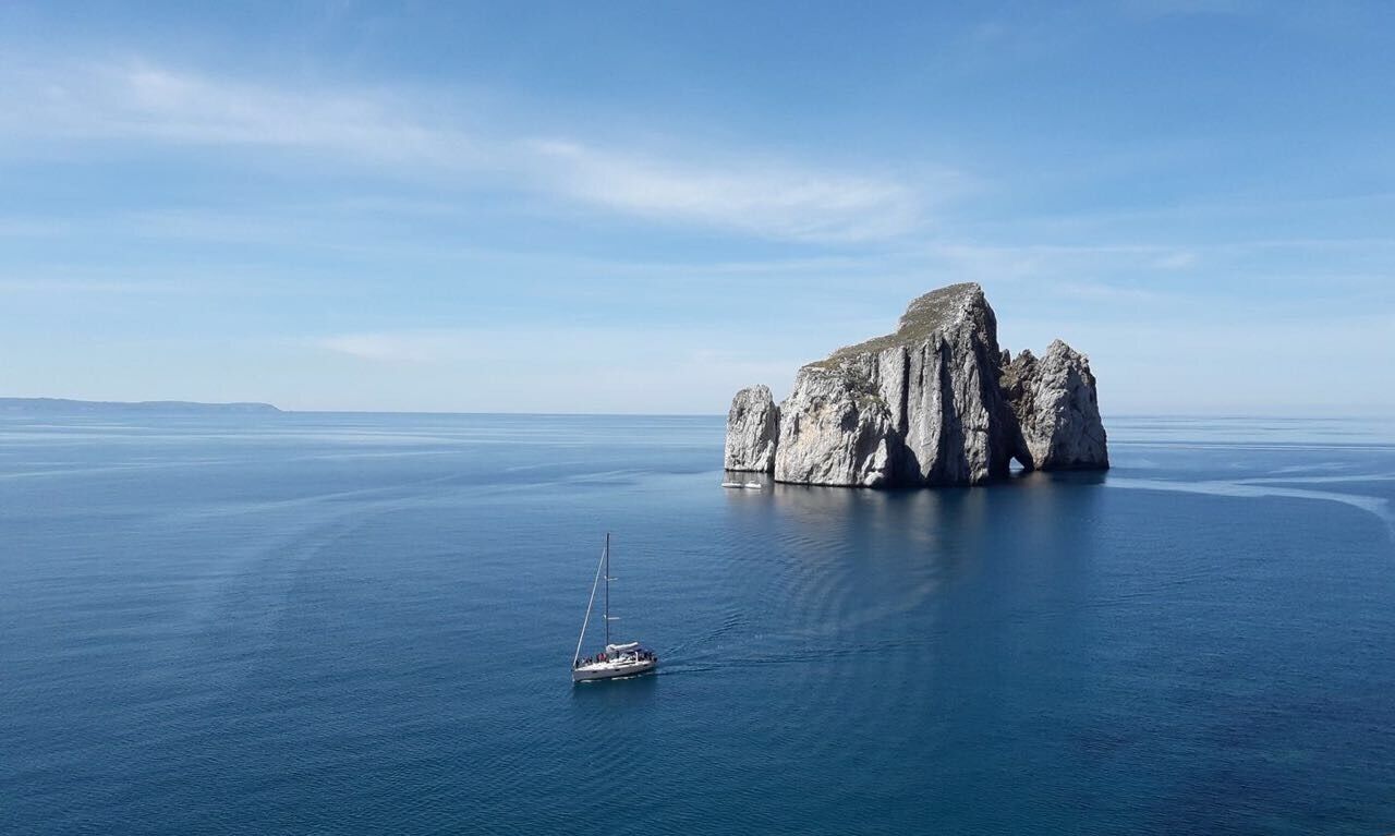 A clear view of a large rock formation surrounded by calm sea, with a sailboat in the foreground.