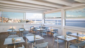 A restaurant with tables and chairs arranged on a terrace with a view of the sea and cityscape.