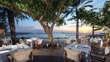 An outdoor dining area with tables set for two, overlooking a body of water at sunset.