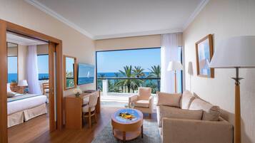 A hotel room with a bed, a desk, a sofa, a round coffee table with fruit and wine, and a balcony with a view of the ocean and palm trees.