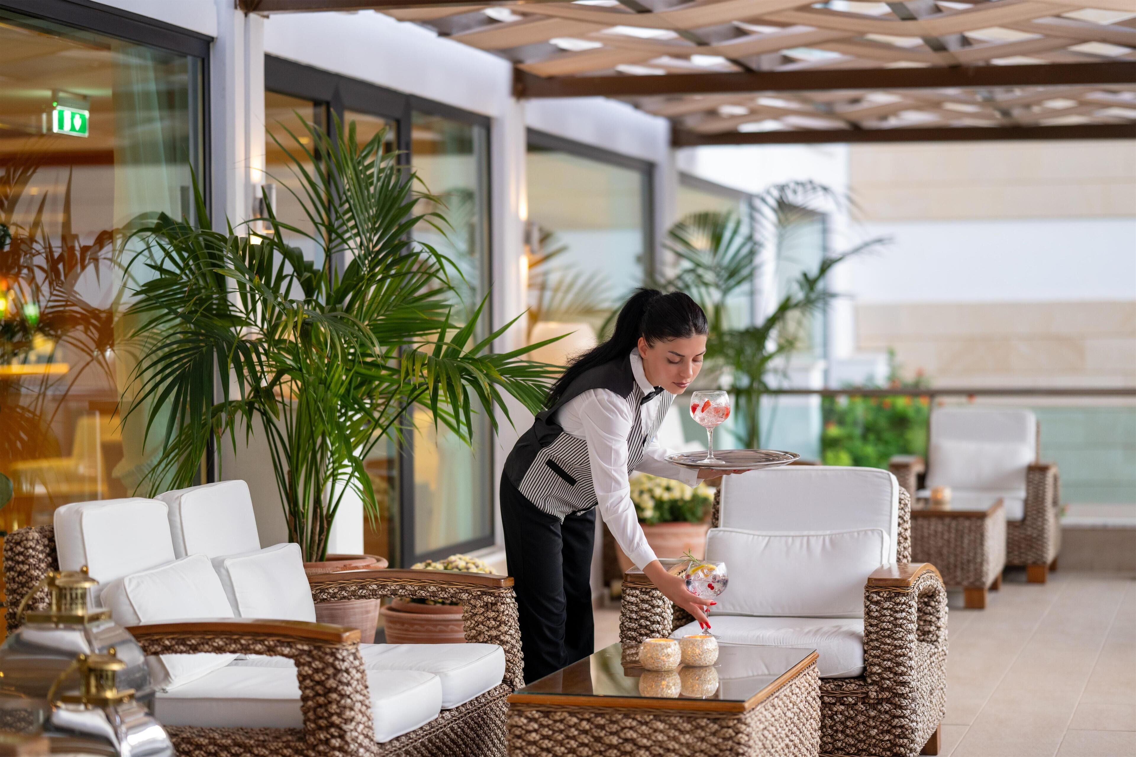 Vestíbulo de hotel con muebles de mimbre, un camarero sirviendo una bebida y grandes plantas en macetas.