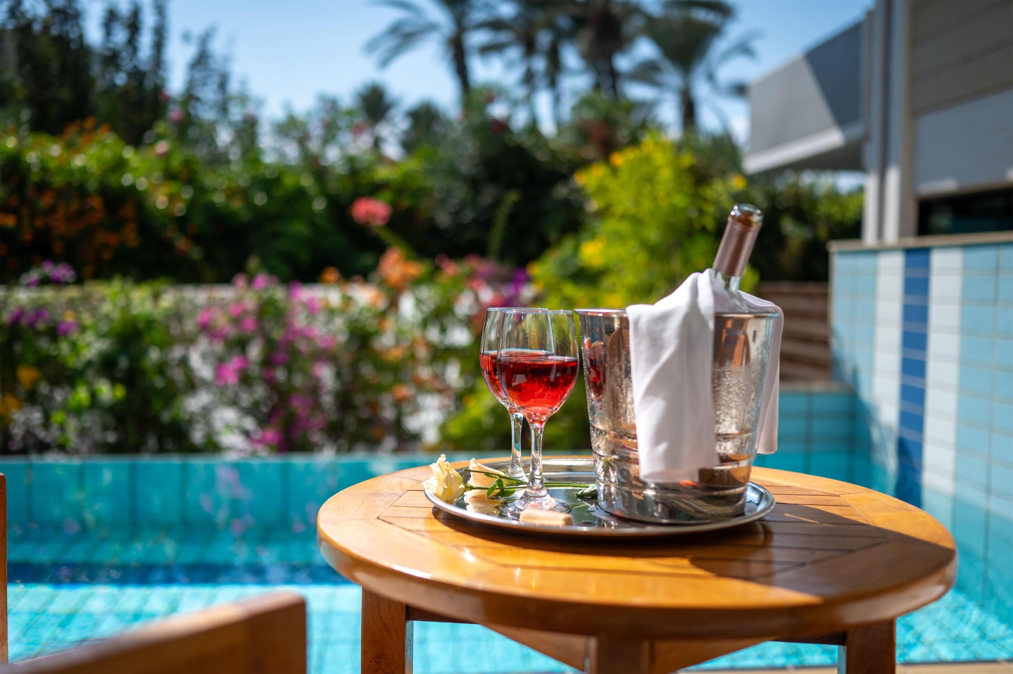 A table with a bottle of red wine, a glass of red wine, and a glass of water by a poolside.