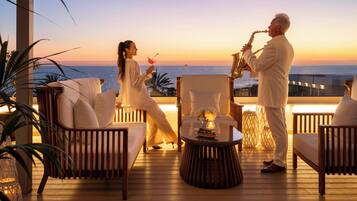 Una pareja en un balcón con vistas al océano, un saxofonista tocando y una mesa con una vela.