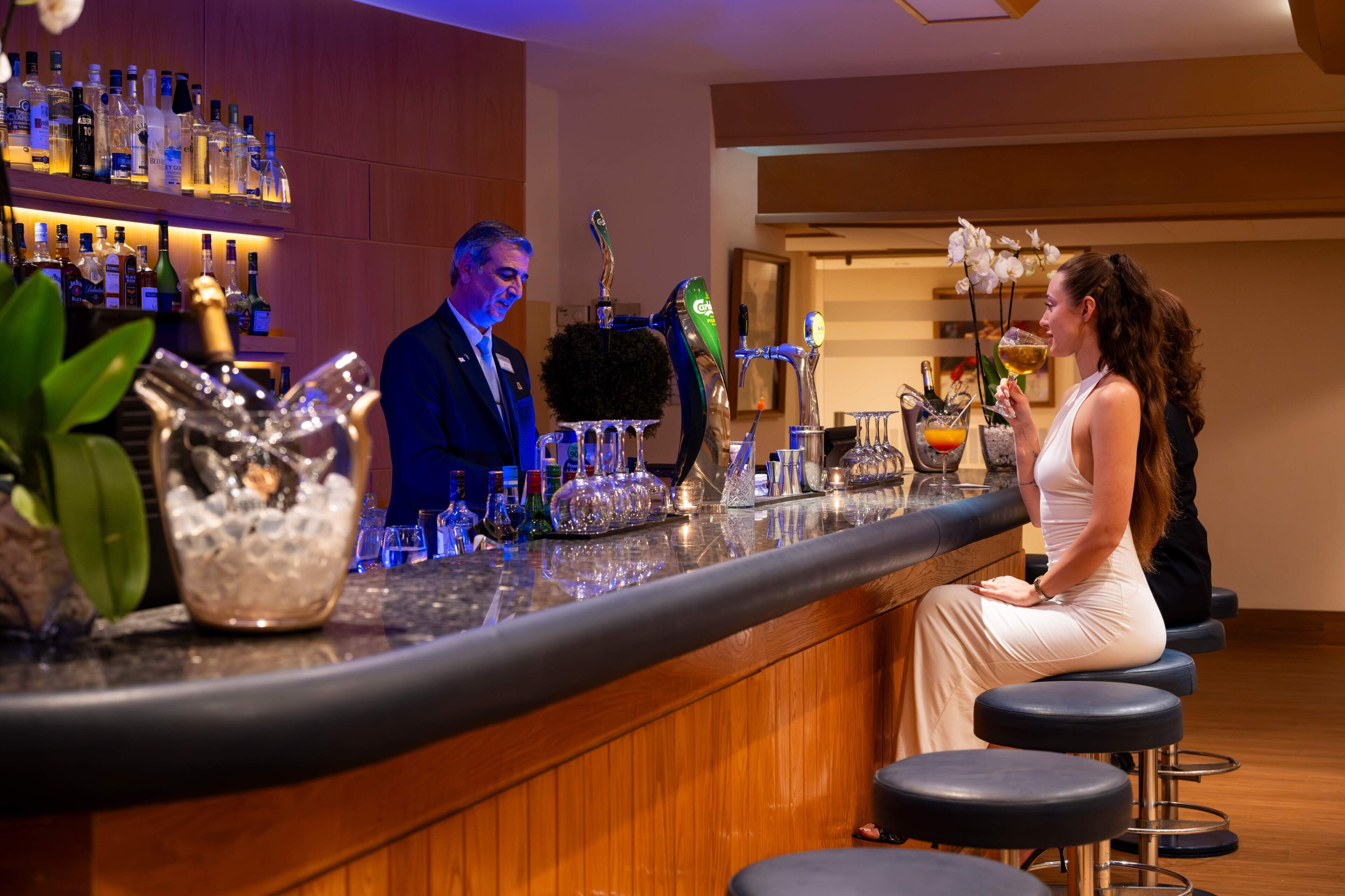 Un espace bar avec un barman, une femme assise et une variété de bouteilles et de verres sur le comptoir.
