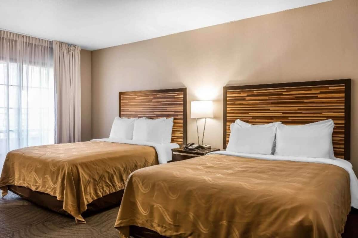 A hotel room with two beds, a bedside table with a lamp, and a window with curtains.