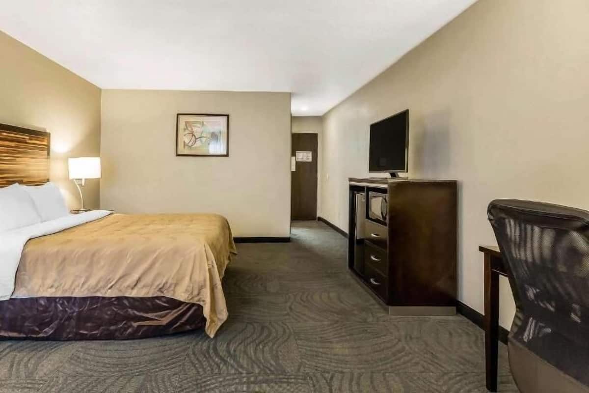 A hotel room with a bed, a desk with a lamp, a dresser, a television, and a painting on the wall.