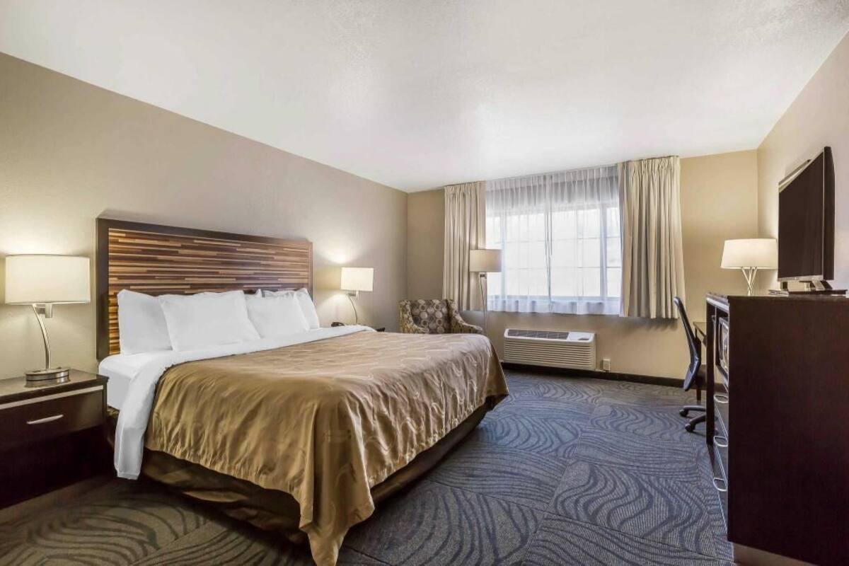 A hotel room with a large bed, two bedside tables with lamps, a desk with a chair, a TV, and a window with curtains.