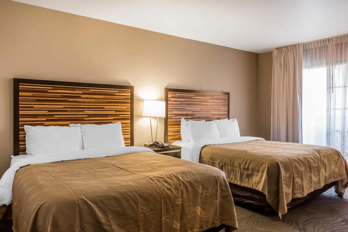 A hotel room with two beds, wooden headboards, white bedding, a bedside table with a lamp, and a window with curtains.