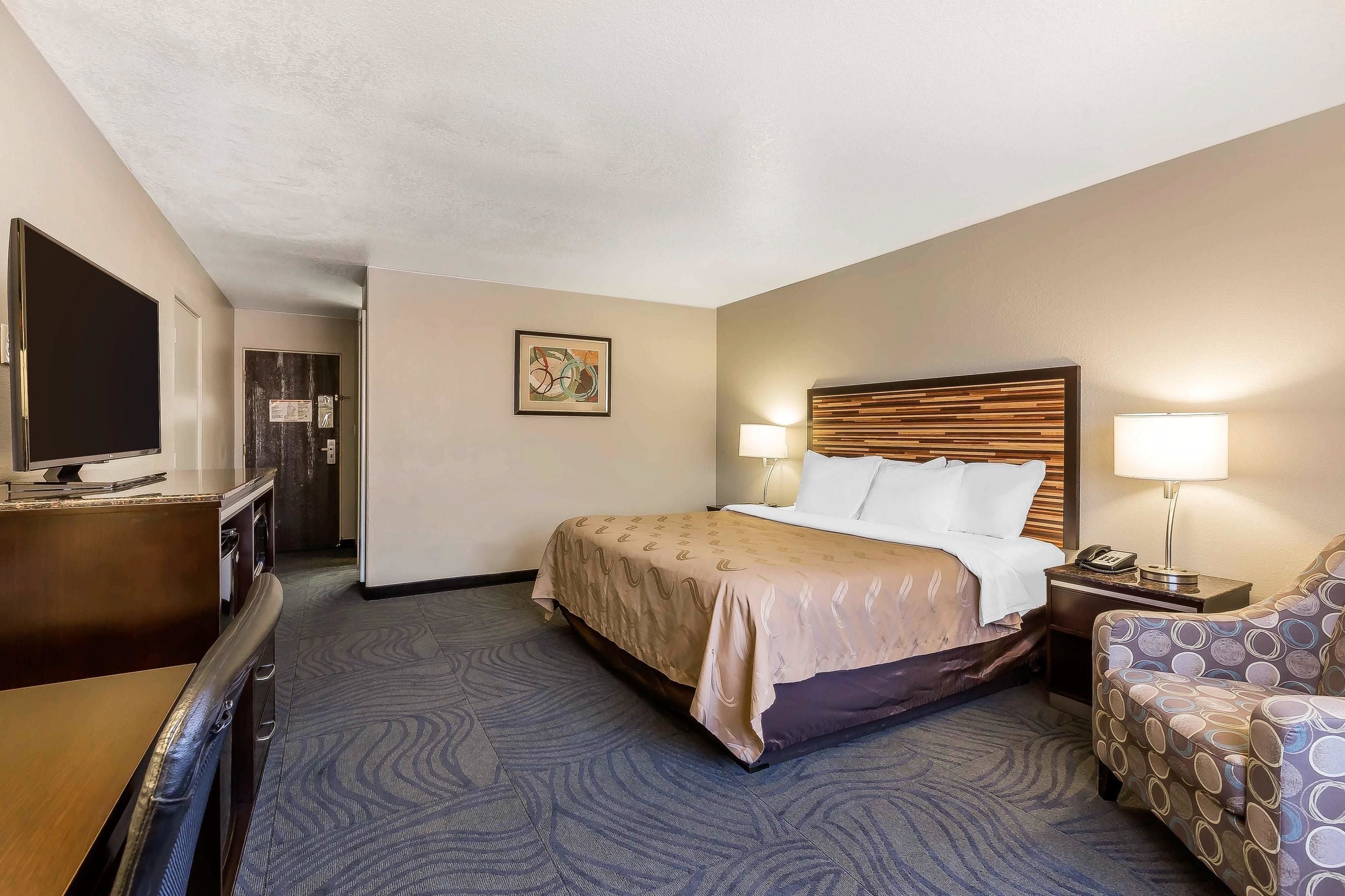 A hotel room with a bed, a desk with a television, a chair, and a nightstand with a lamp.