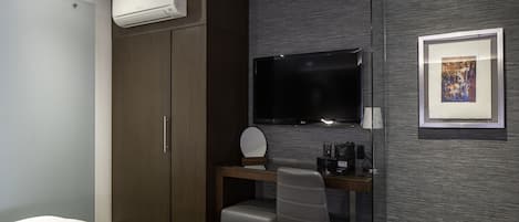 A modern hotel room with a bed, a desk with a chair, a flat-screen TV, an air conditioning unit, and a framed picture on the wall.