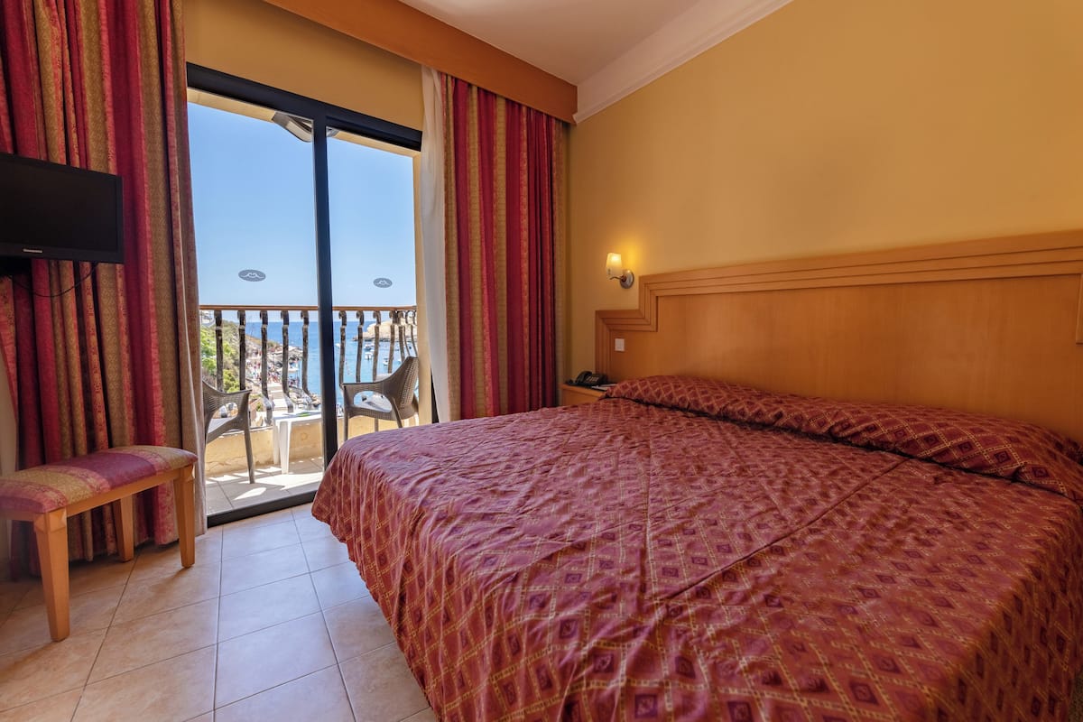 Habitación de hotel con una cama grande, un banco, un televisor y un balcón con vistas al mar.