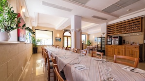 Uma longa mesa de jantar posta com cadeiras, um suporte para vinhos e uma máquina de café em um ambiente bem iluminado com janelas amplas.