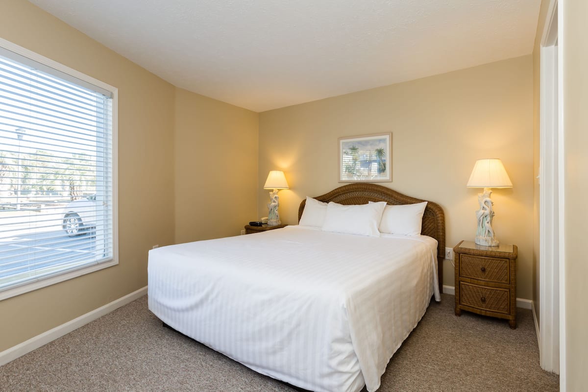 A neatly made bed with white linens in a room with beige walls, a window with blinds, and two bedside lamps.