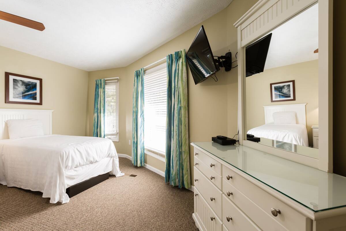 A bedroom with a bed, a dresser, a television mounted on the wall, and a window with curtains.