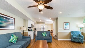 A living room with a ceiling fan, wooden coffee table, two sofas with patterned cushions, a blue armchair, and a kitchen in the background.