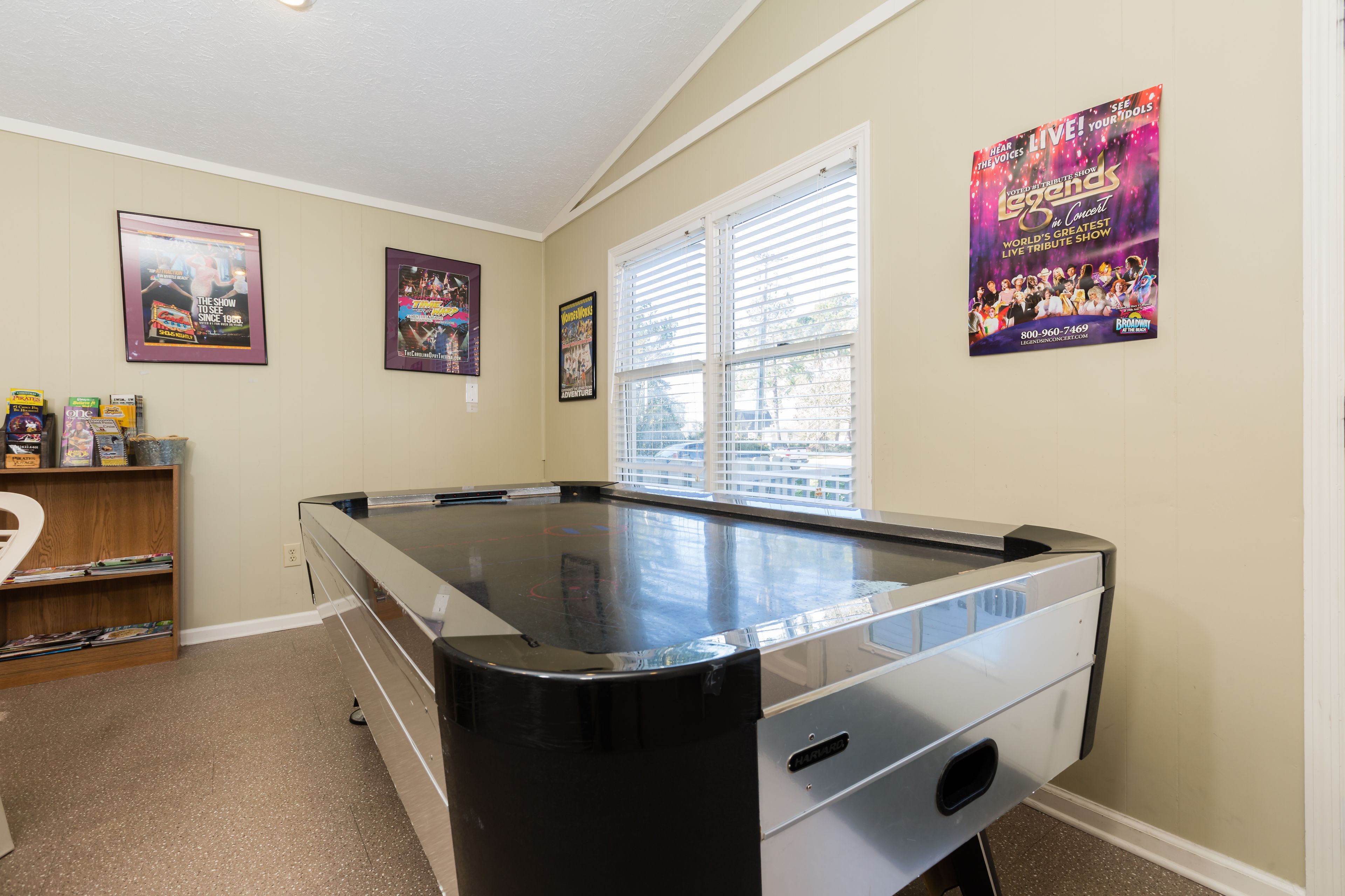Une table de billard dans une pièce aux murs de couleur beige, avec une fenêtre équipée de stores et des affiches accrochées au mur.