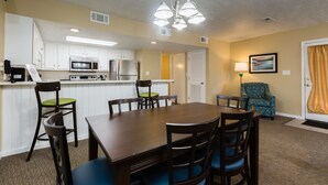 A compact kitchen with white cabinets, a microwave, and a refrigerator. There is a dining table with chairs and a blue armchair.