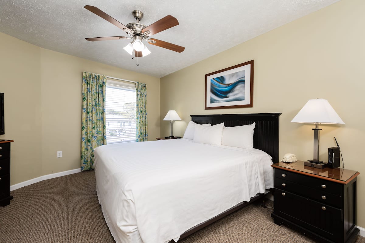 A hotel room with a bed, a nightstand, a lamp, a ceiling fan, and a window with curtains.