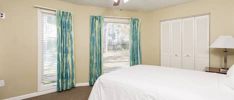 Un dormitorio con cama, ventilador de techo, ventana con cortinas, mesita de noche y armario.